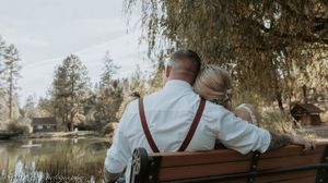 A panoramic view of a wooden bench under willow trees beside a tranquil pond in a peaceful park setting.
