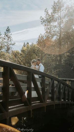 A couple shares a romantic moment on a wooden bridge surrounded by sunlit trees in an outdoor setting.