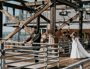 A couple in wedding attire stands on opposite sides of a rustic wooden deck with industrial architecture.