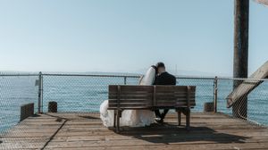 A panoramic view of a wooden pier extending into calm blue waters with benches and people relaxing on a sunny day.