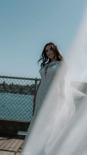 A white wedding dress with lace details photographed against a bright blue sky and waterfront backdrop.