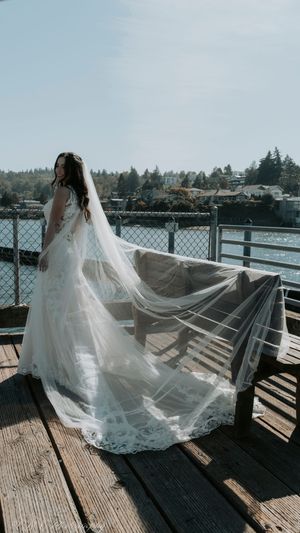 A flowing white wedding dress and veil catch the breeze on a waterfront deck with scenic lake views in the background.