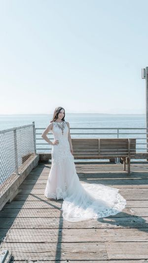 A bride in a flowing white lace gown poses gracefully on a wooden pier overlooking the ocean on her wedding day.