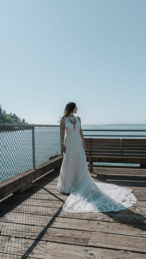 A wedding dress train trails along a wooden pier overlooking the ocean on a sunny day.