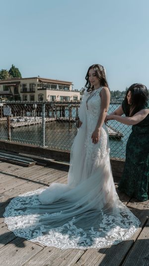 Wedding dress train spreads beautifully across wooden boardwalk as bride gets final adjustments near waterfront.