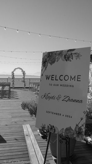 Wedding welcome signs displayed on a wooden deck with string lights and ocean views at sunset.