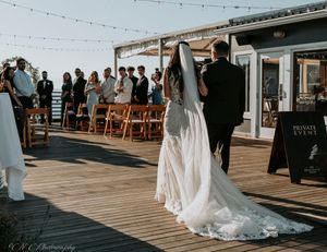A wedding ceremony takes place on a wooden deck adorned with string lights as guests watch from wooden chairs.