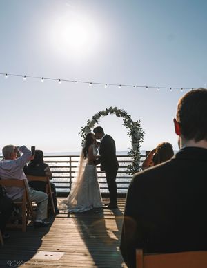 Silhouettes of wedding guests seated on a pier during an outdoor ceremony at sunset.