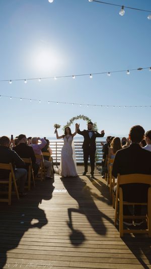Wedding ceremony on a wooden deck overlooking the ocean with string lights and guests seated in wooden chairs at sunset.