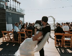 A wedding celebration on a waterfront deck with wooden chairs and string lights overhead.