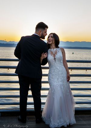 A romantic sunset wedding photo of a couple in formal attire standing on a pier overlooking the water.
