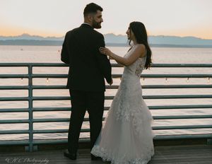 A couple in wedding attire shares a romantic moment on a pier at sunset with mountains in the background.