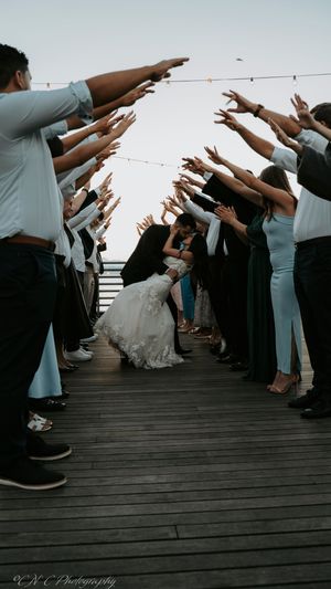 Wedding guests form a tunnel with raised arms on wooden boardwalk during sunset celebration.