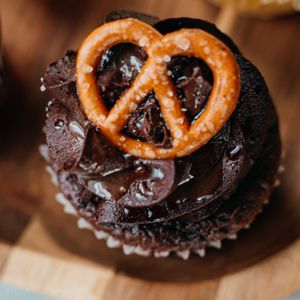 A chocolate cupcake topped with dark chocolate frosting and decorated with a salted pretzel twist on top.