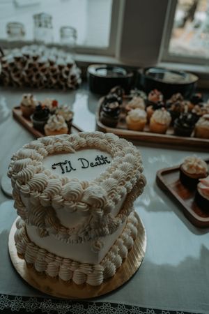 A white wedding cake decorated with piped frosting swirls and 'Til Death' written on top sits next to chocolate cupcakes.