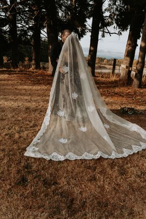 A long cathedral wedding veil with lace appliques trails on autumn ground in a forest setting.