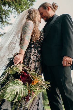 Couple in black wedding attire share intimate moments while holding a lush bouquet with red flowers and greenery.