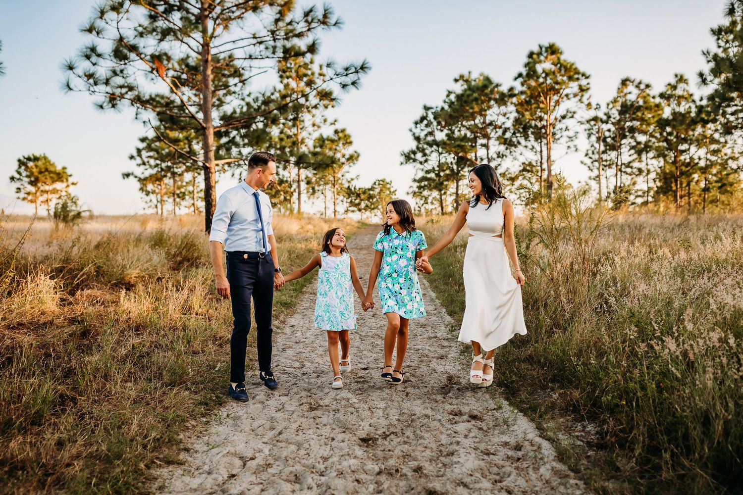 A family walks together on a dirt path through tall grass and pine trees at golden hour in natural lighting.