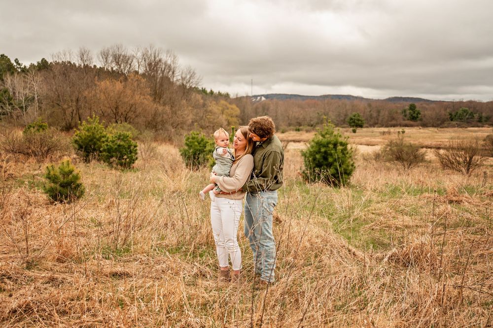 A couple embraces in a rustic autumn field with rolling hills and cloudy skies in the background.