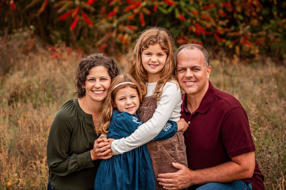 A family wearing coordinated navy blue and burgundy outfits poses together in a fall field with autumn foliage.