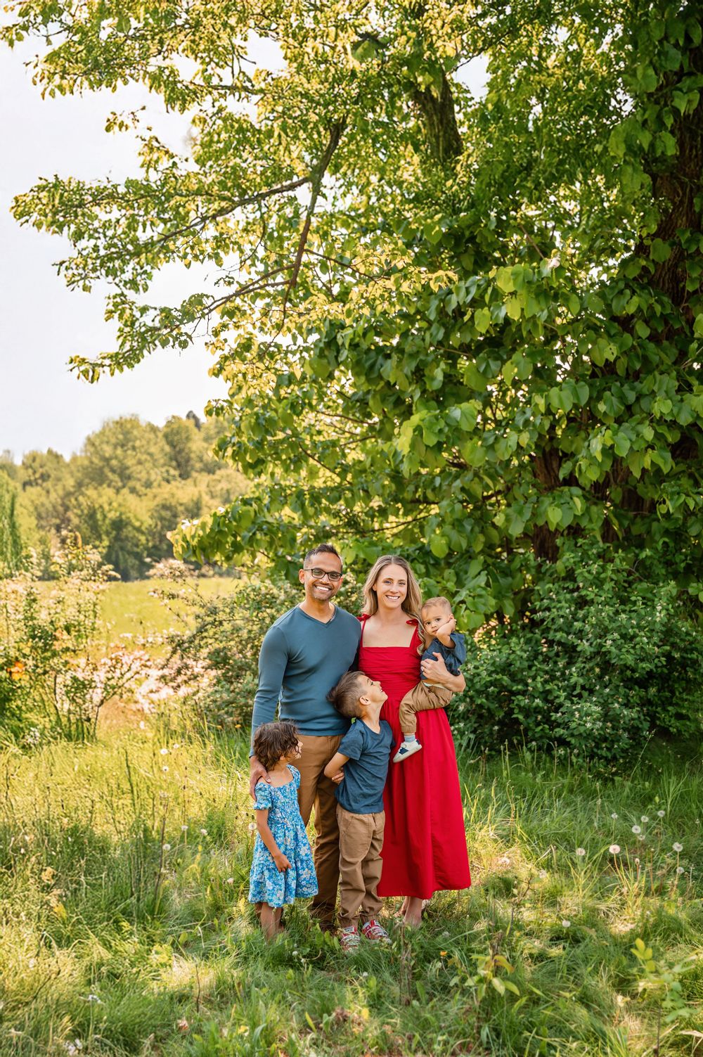 A family in casual attire poses together outdoors under green trees on a sunny summer day in a grassy field.