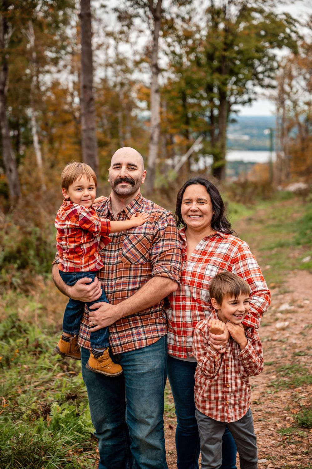 A family wearing coordinating plaid and flannel shirts poses together on a wooded autumn trail.