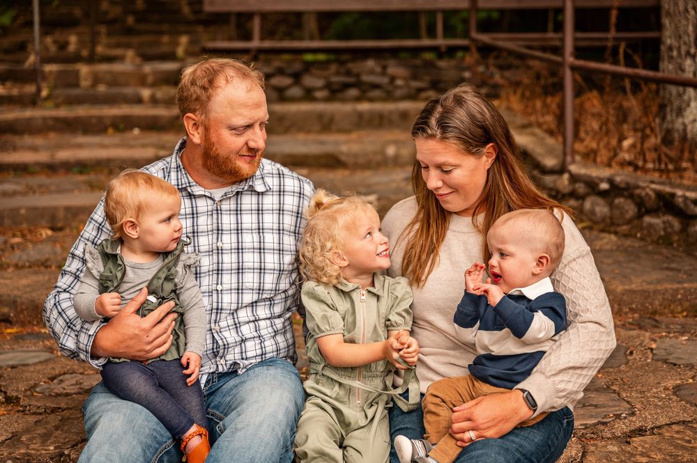 A family sits together on fallen autumn leaves during an outdoor photo session in a rustic setting.