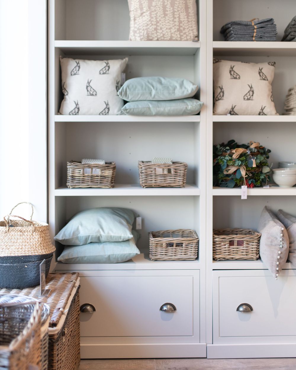 White built-in storage unit with shelves displaying woven baskets, pillows and decorative items in a neutral color scheme.