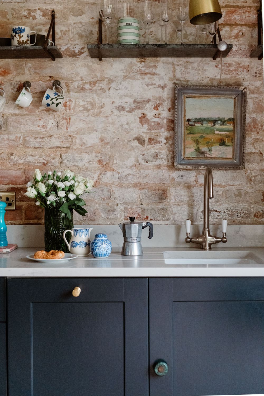 Modern kitchen interior with navy blue cabinets, exposed brick wall, and brass pendant light fixture above marble countertop.