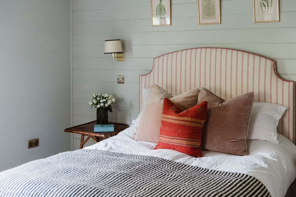 A bedroom with pink and white striped wallpaper features a curved headboard, white bedding, and coral accent pillows.