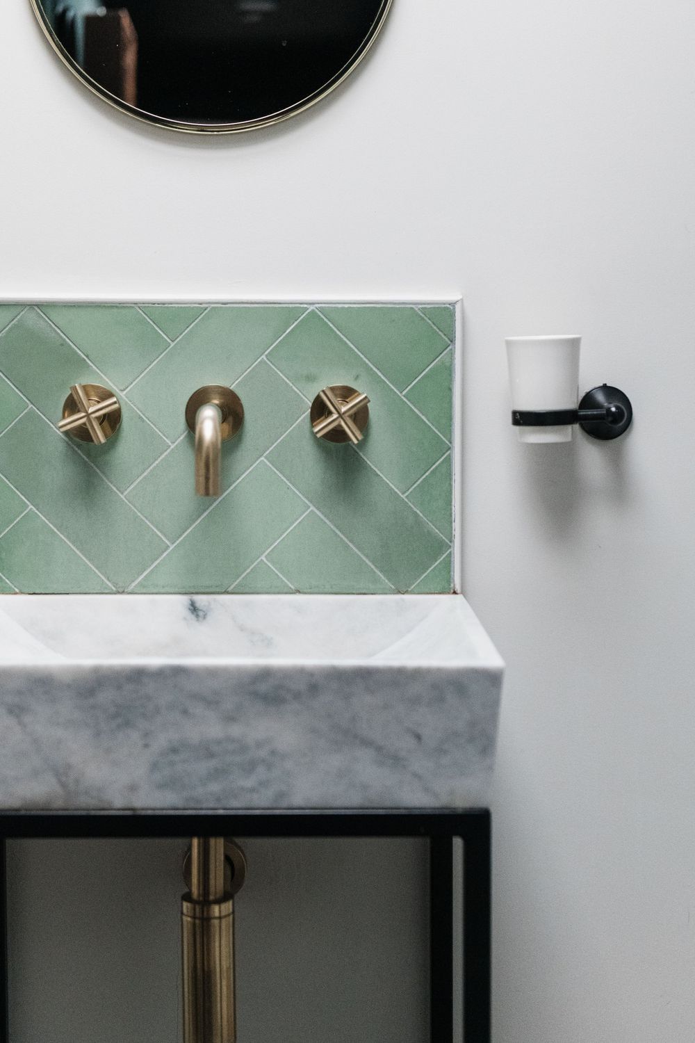 Modern bathroom interior with mint green diagonal tile backsplash, round mirrors, and black and brass hardware accents.
