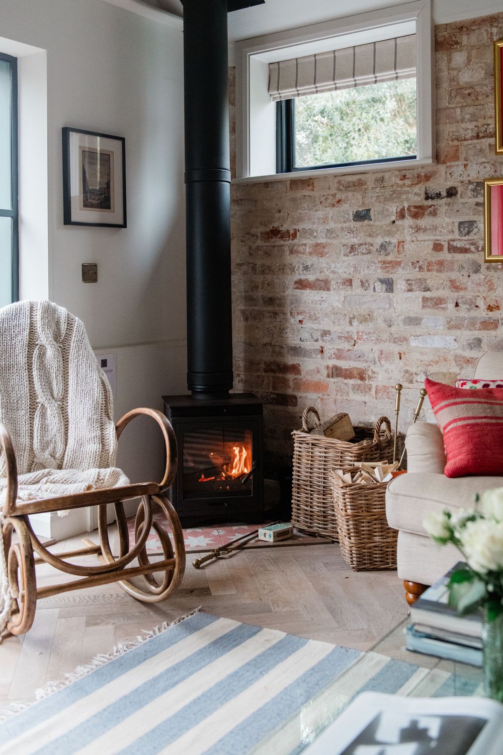Cozy living room corner with wood burning stove, rocking chair, exposed brick wall and striped blue rug.