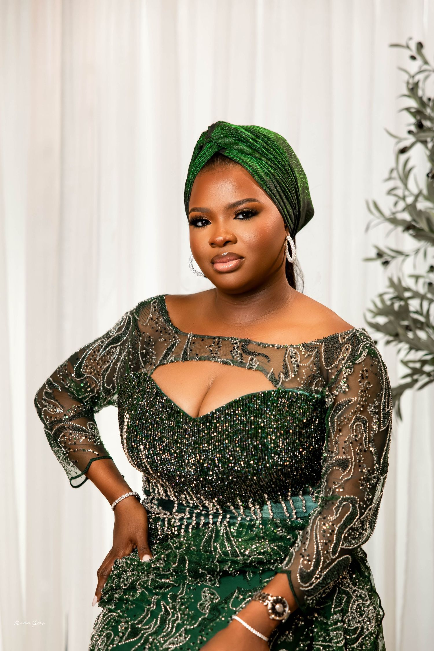 Elegant emerald green evening gown with intricate beading and matching head wrap against light curtained backdrop.