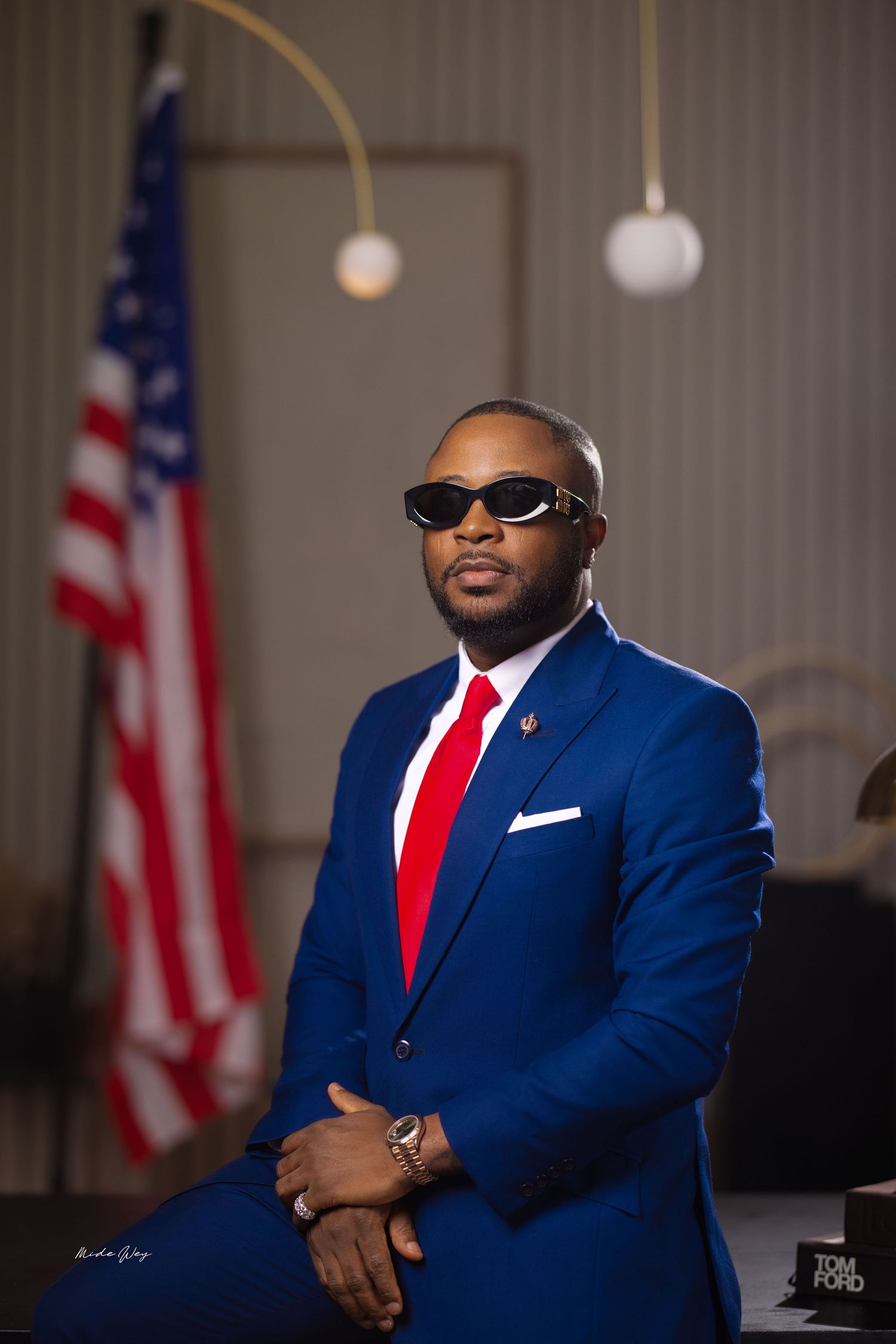Professional wearing a navy blue suit and red tie poses confidently in front of American flags.