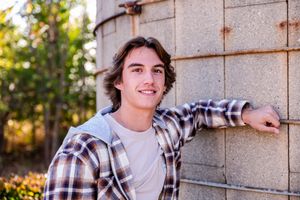 A person in a plaid flannel shirt and white t-shirt poses against a concrete wall in warm sunlight.