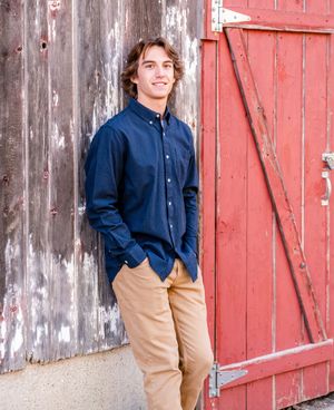 Casual outdoor portrait against rustic barn wall with red door showing navy shirt and khaki pants.