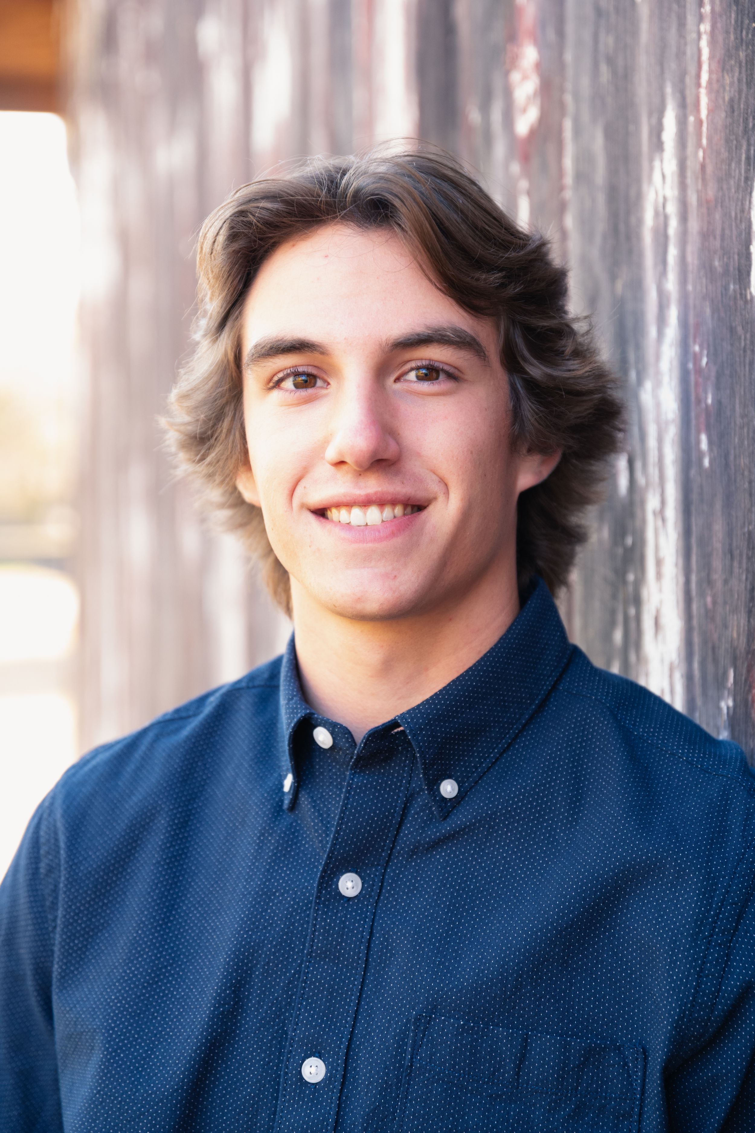 A person wearing a navy blue button-down shirt smiles warmly against a rustic wooden background.