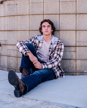 Someone in a plaid flannel shirt and jeans sits casually against a textured concrete wall outdoors.