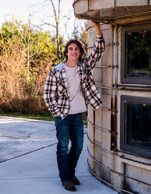 Person in plaid shirt and jeans sits casually on concrete ledge against stone wall in outdoor setting at sunset.