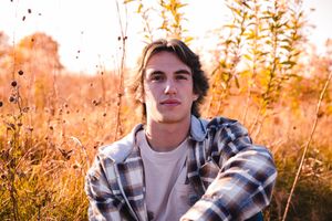 A person wearing a plaid flannel shirt poses in golden autumn sunlight amidst tall grass and foliage.