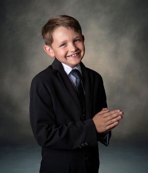 A young student in formal attire with a black cardigan and tie poses for a portrait against a dark studio backdrop.