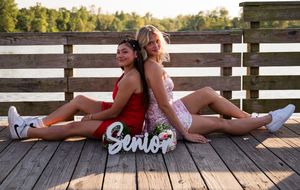 Two students in casual attire sit back-to-back on a wooden deck with a decorative 'Senior' sign between them.