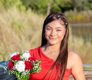 A person in a bright red dress poses outdoors with a bouquet of red and white flowers near a lake.