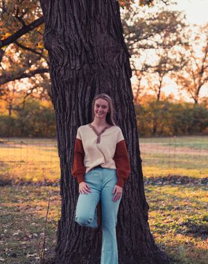 Person in light colored top and jeans leaning against large tree trunk at sunset in autumn park setting.