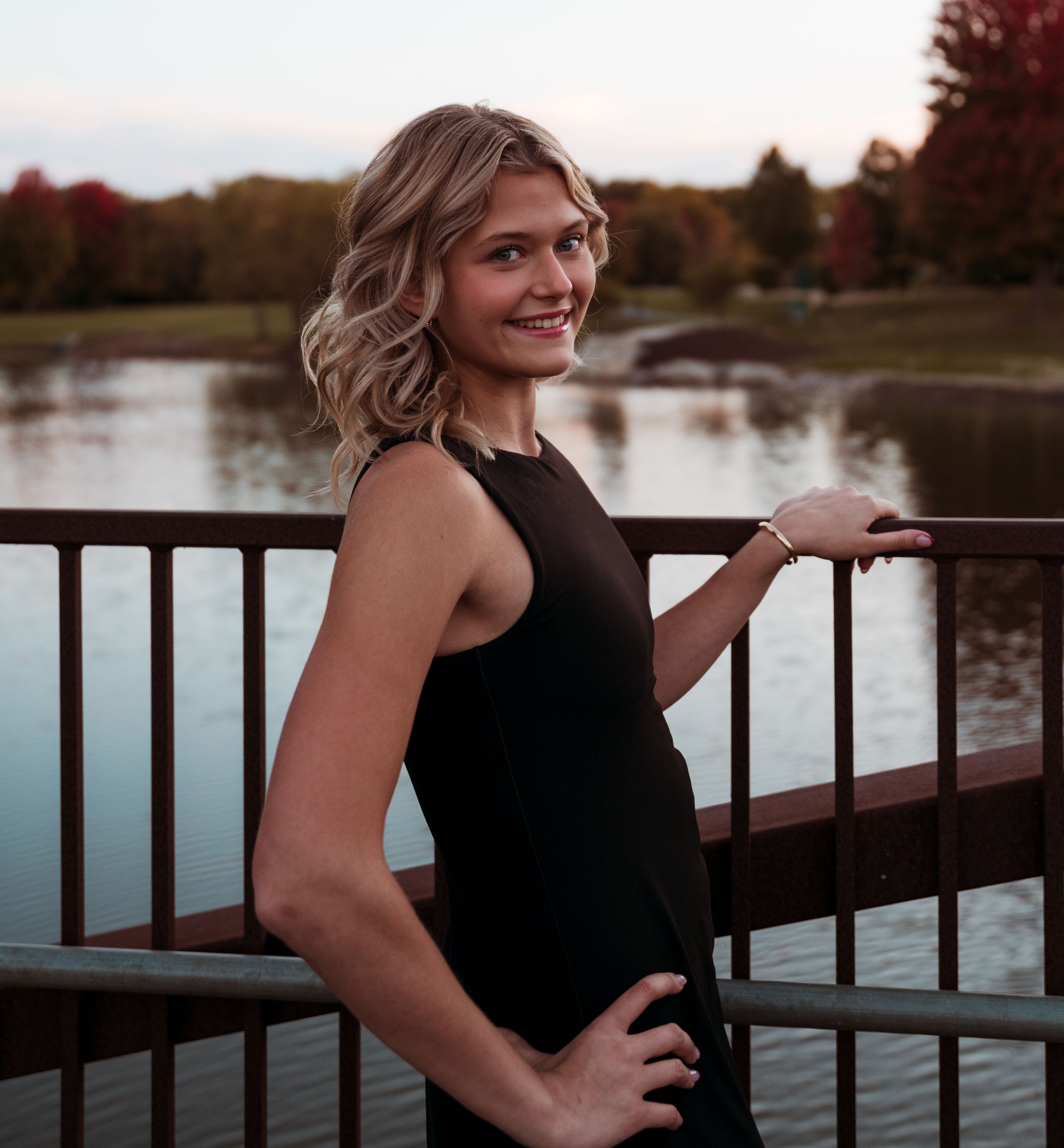 A person in a black sleeveless dress poses on a bridge railing at sunset with autumn trees and a lake in the background.