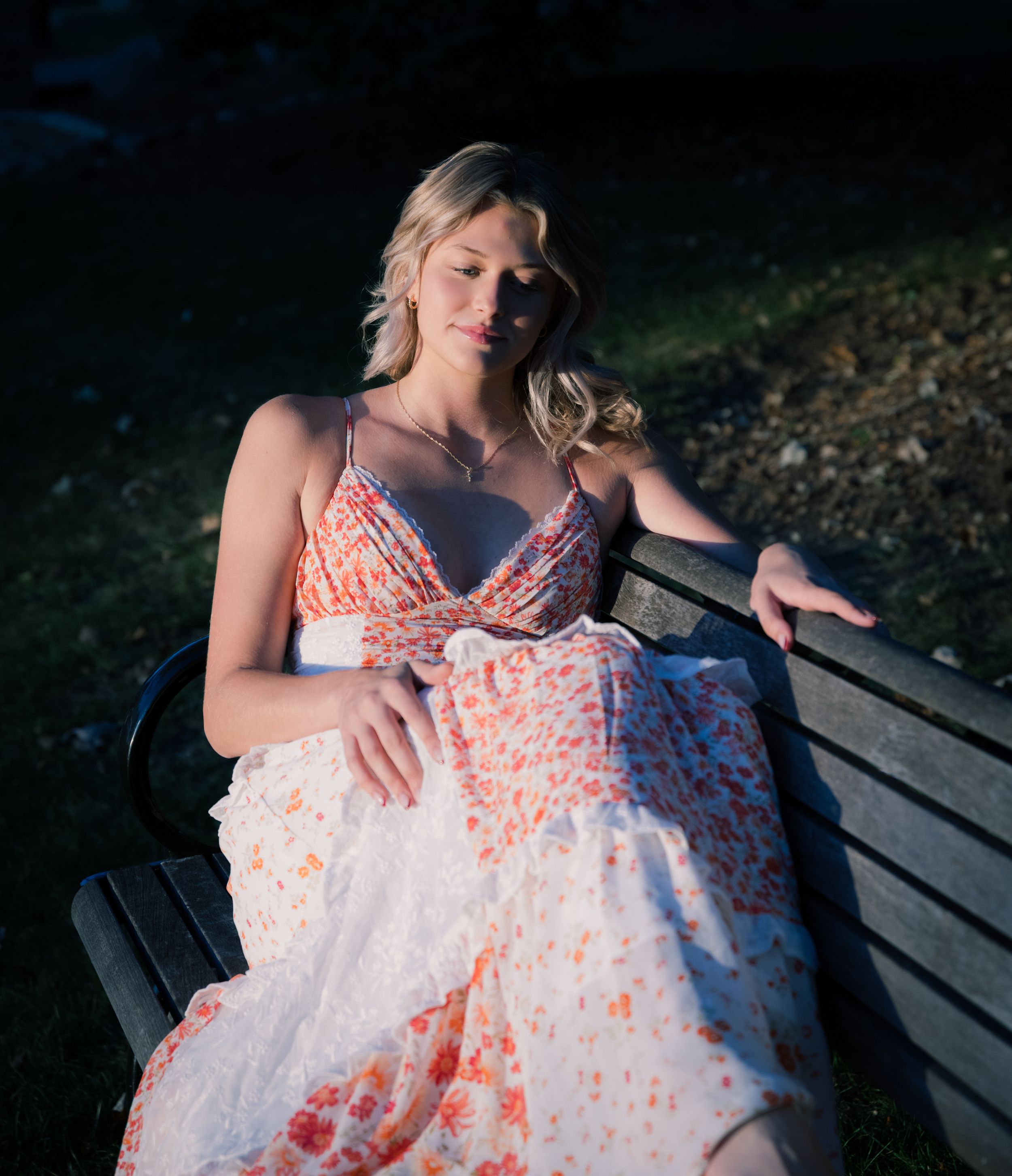 A person wearing a white and coral floral dress sits on a wooden bench in a moody outdoor setting.