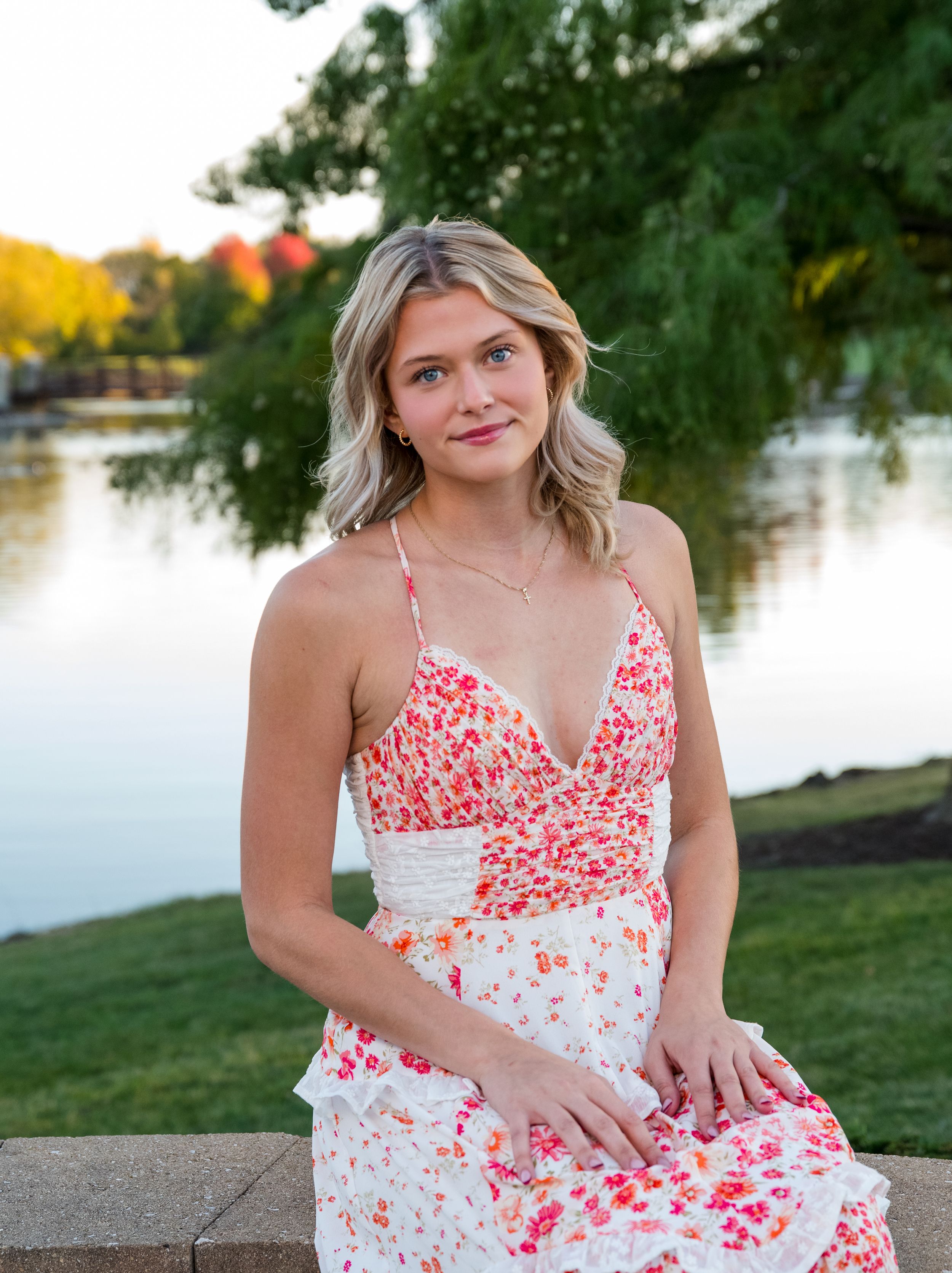 A woman in a floral white and red sundress poses gracefully by a lake during golden hour.