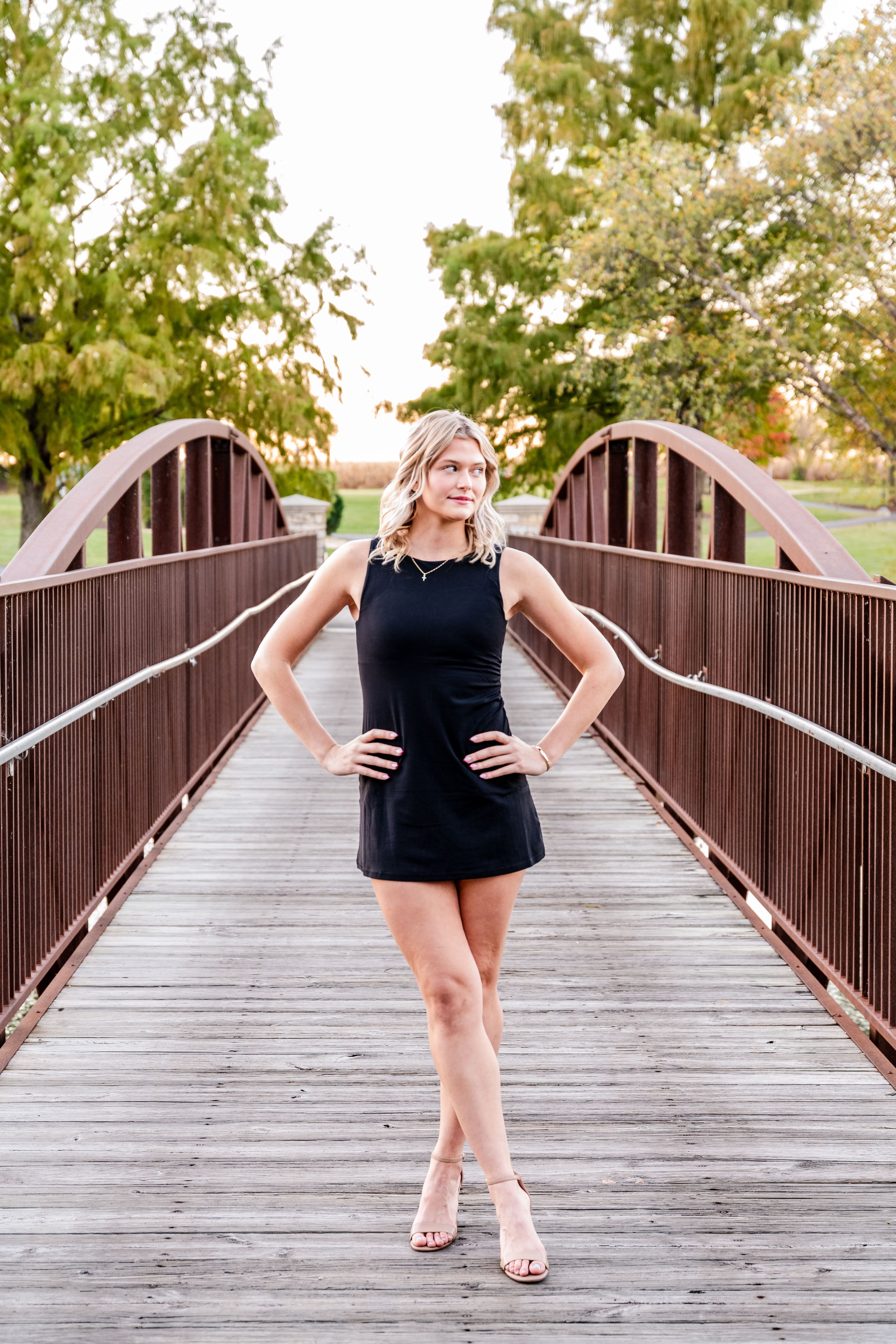 A black mini dress and heels outfit photographed on a wooden bridge with greenery in the background.