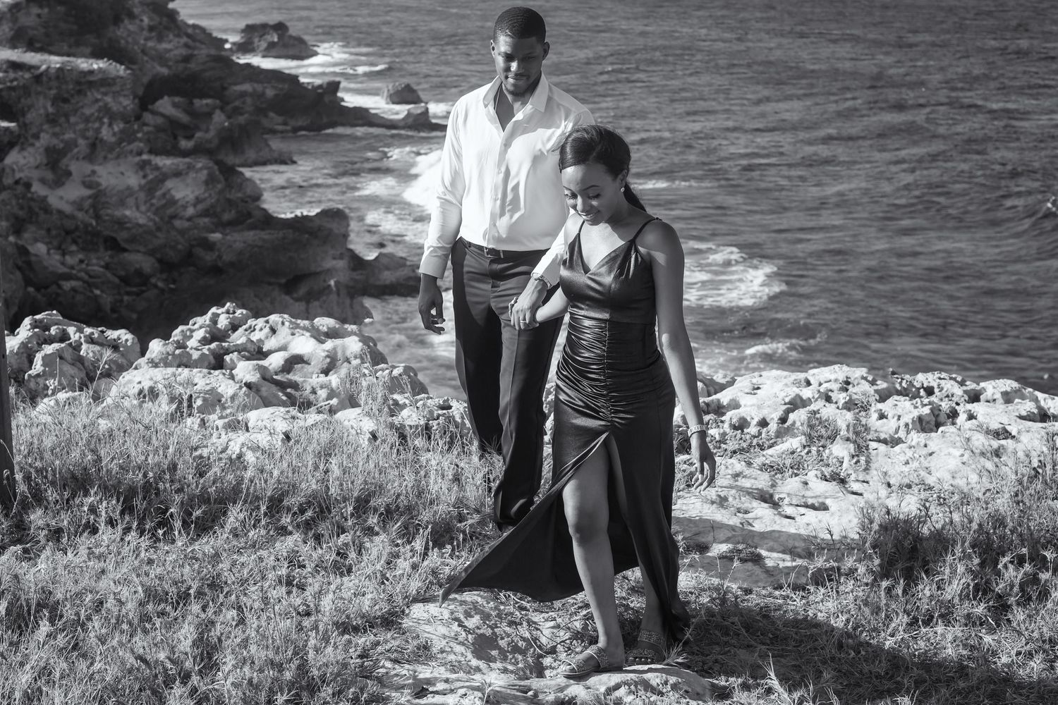 A black and white photograph of two people standing near a rocky coastal cliff with ocean waves crashing below.