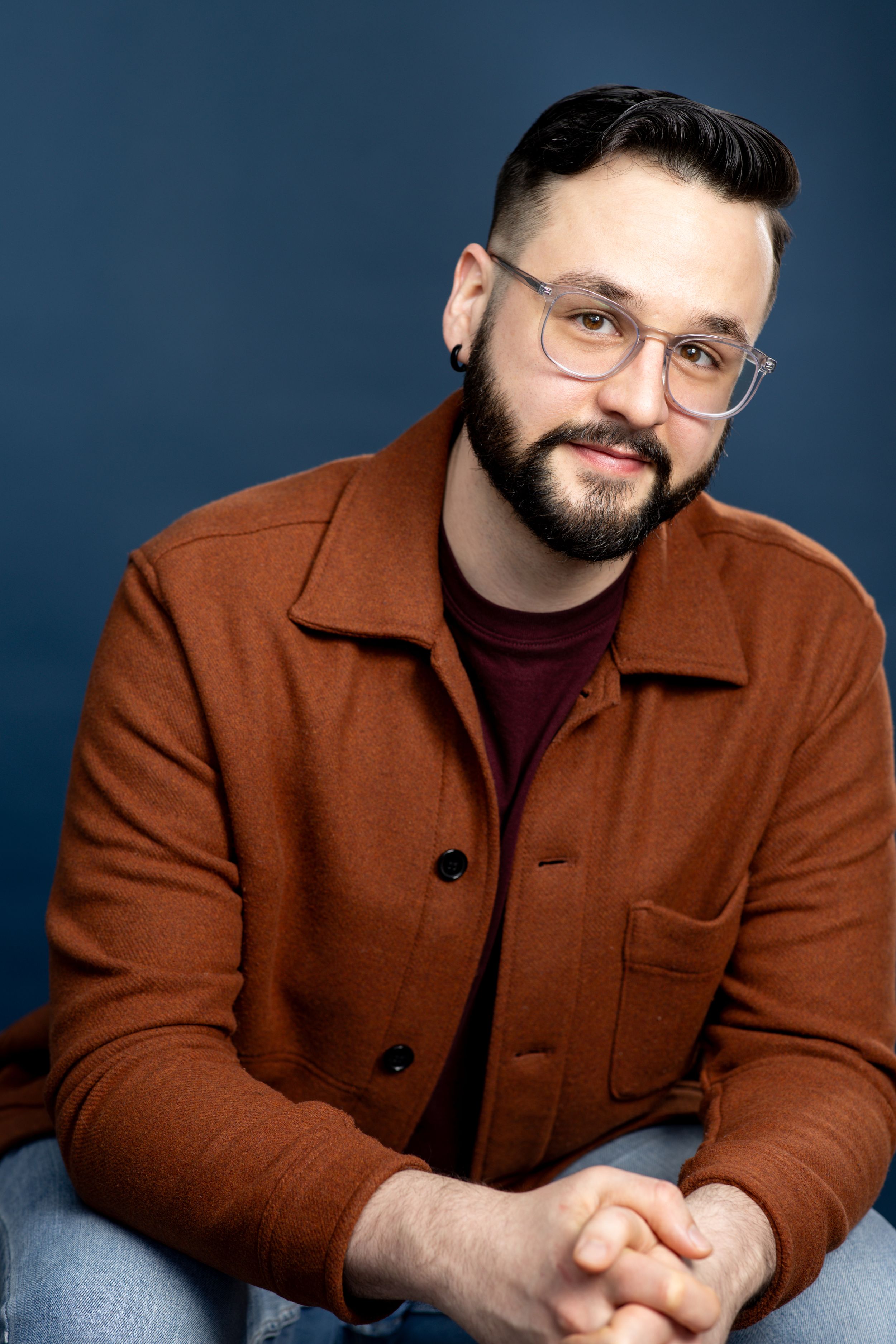 A headshot of a man with glasses wearing orange jacket against a dark blue background. Twin cities headshots. Theatre headshots. Headshots.